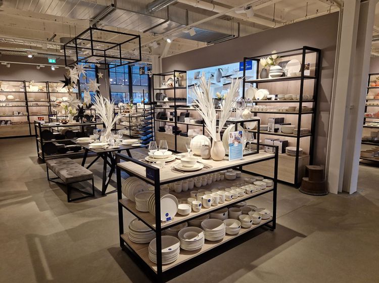 Im Villeroy&Boch Outlet Mettlach stehen in einem modernen Geschäft übersichtlich angeordnete Regale mit weißem Geschirr, Schalen und Küchenutensilien. Auf den zentralen Tischen stehen dekorative Vasen mit weißem Laub. Der Raum ist gut beleuchtet und sauber und übersichtlich gestaltet.