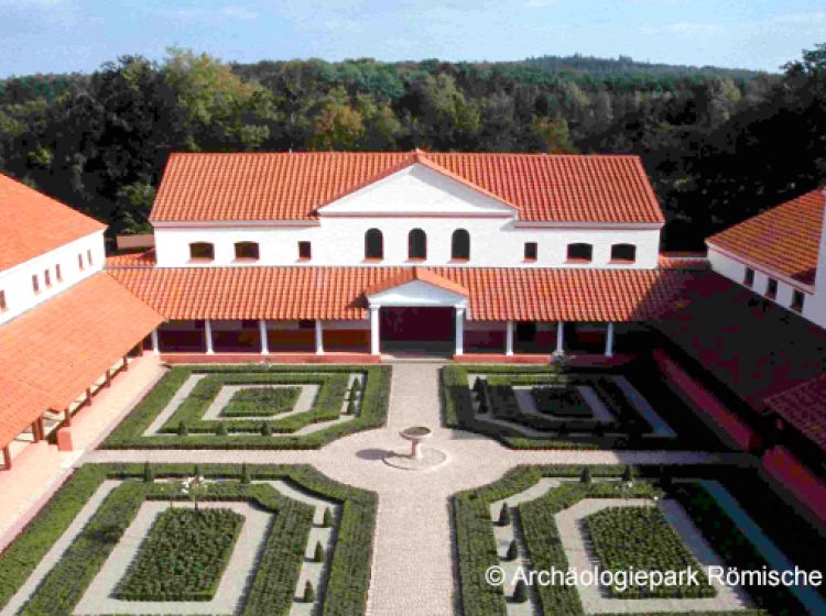 Eine rekonstruierte römische Villa mit roten Ziegeldächern umgibt einen zentralen Hof mit ordentlich geschnittenen Hecken, Kieswegen und einem runden Brunnen; im Hintergrund sind Bäume und Wald zu sehen (Archäologiepark Römische Villa Borg).