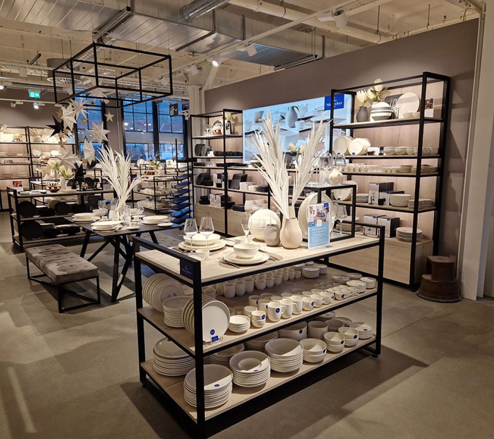 Im Villeroy&Boch Outlet Mettlach stehen in einem modernen Geschäft übersichtlich angeordnete Regale mit weißem Geschirr, Schalen und Küchenutensilien. Auf den zentralen Tischen stehen dekorative Vasen mit weißem Laub. Der Raum ist gut beleuchtet und sauber und übersichtlich gestaltet.
