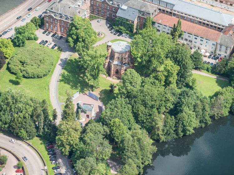 Luftaufnahme eines Parks mit dichten grünen Bäumen, einer gewundenen Straße, geparkten Autos, einem runden Gebäude in der Mitte (Alter Turm, Mettlach) und historischen Backsteinbauten in der Nähe eines Flusses.
